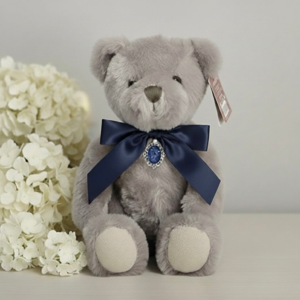 Gray teddy bear with a blue bow and tag with ashes pendant next to white flowers on a light background