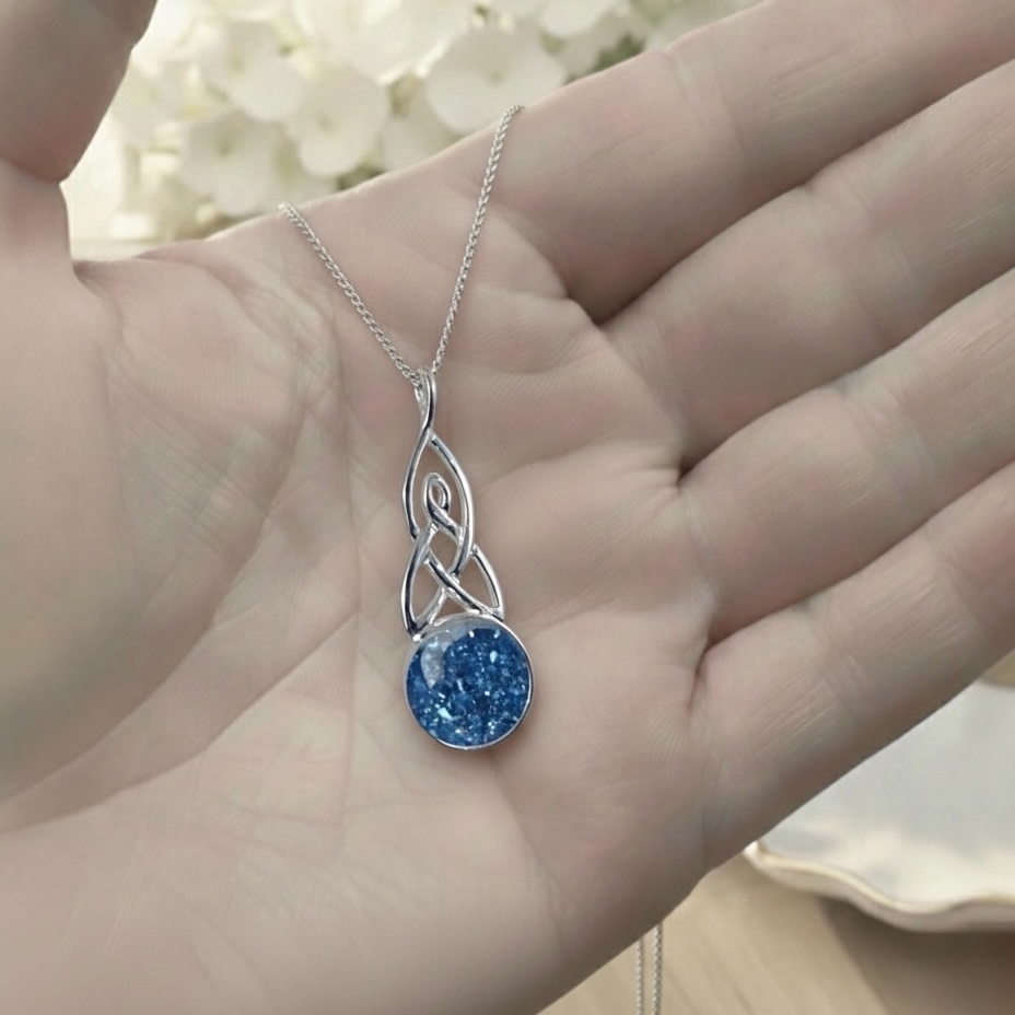 Silver necklace with a blue pendant held in a hand against a blurred floral background
