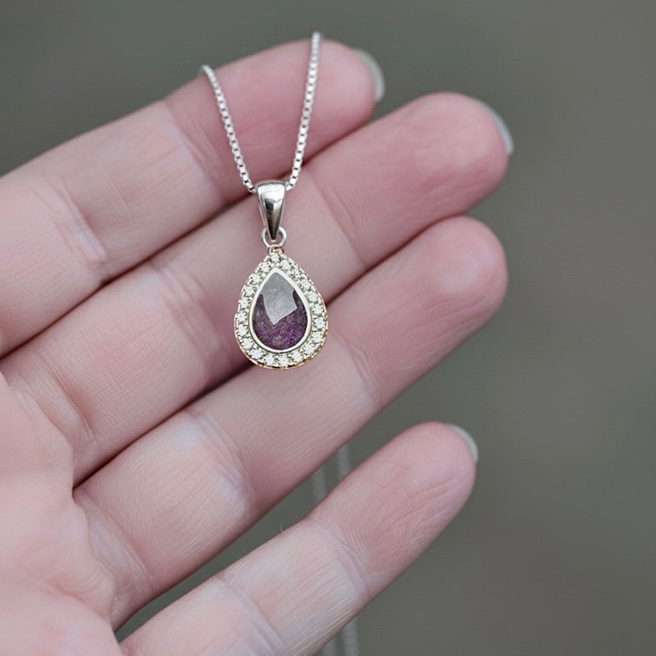 Teardrop-shaped ashes keepsake pendant with purple stone and clear stones on a chain held in a hand.