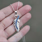 Silver Memory Within feather-shaped ashes keepsake pendant with blue stones on a hand against a neutral background