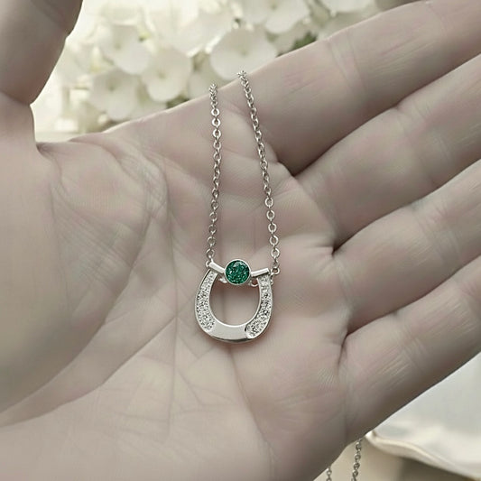 Silver horseshoe necklace with a green ashes gemstone held in a hand against a blurred floral background