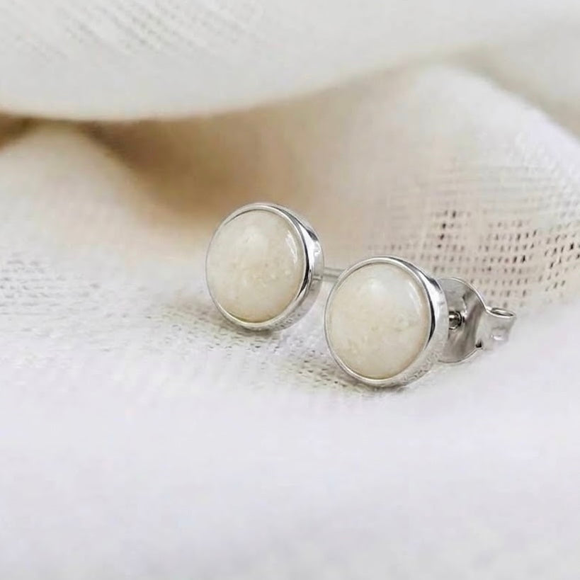 A pair of silver stud earrings with round, breastmilk cabochon gemstones, resting on a soft, textured white fabric background.