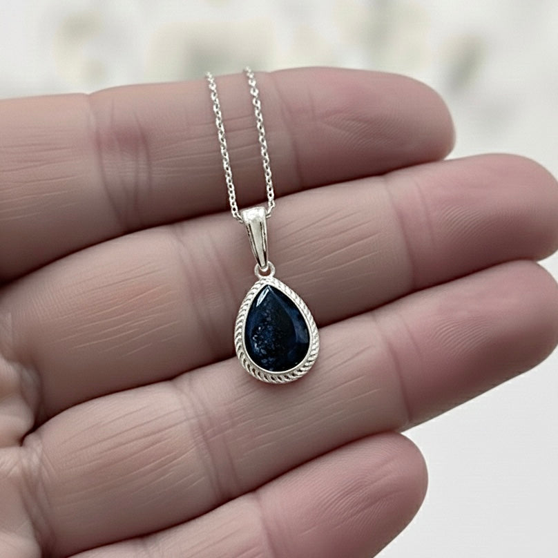 Teardrop-shaped ashes keepsake memorial pendant with black stone on a silver chain held in hand against a blurred background