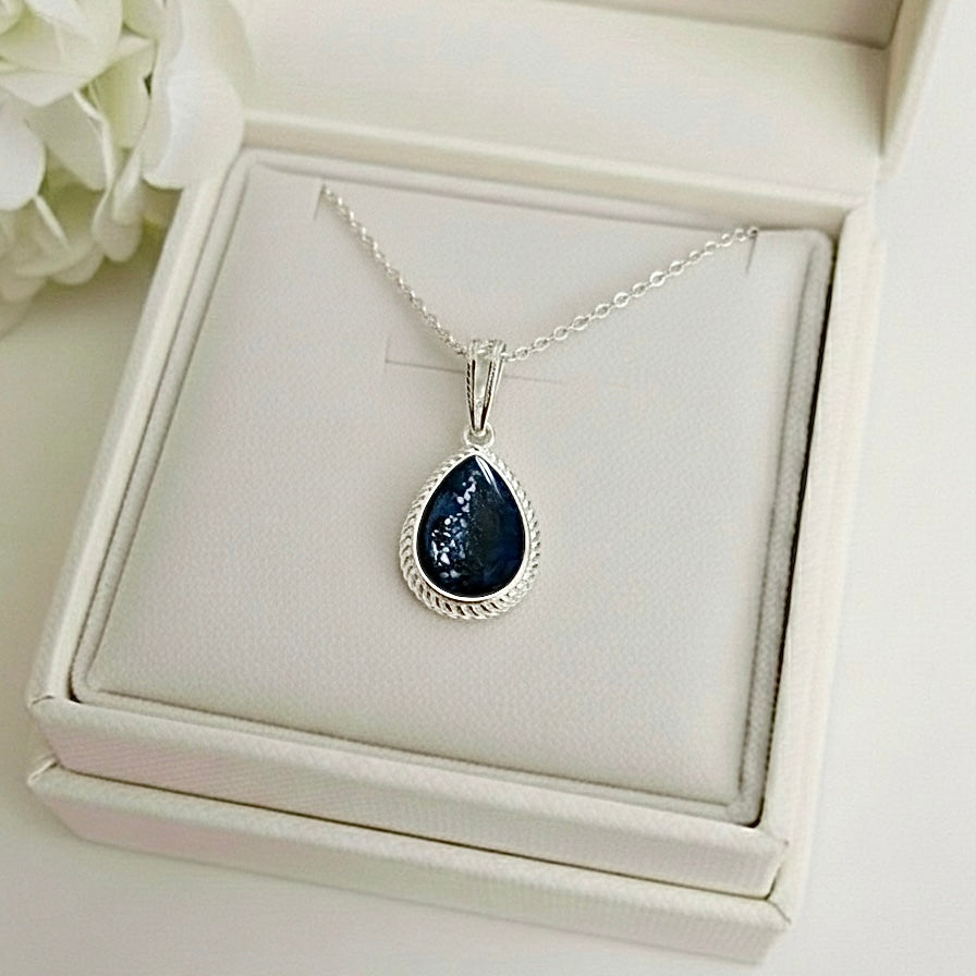 Rope edge Teardrop-shaped ashes keepsake memorial pendant necklace on a memory within white box with flowers in the background