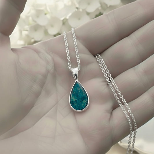Teardrop-shaped ashes keepsake pendant on a silver chain held in a hand with a blurred floral background